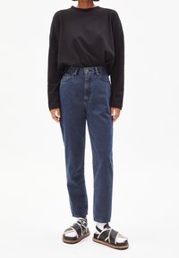 Dunkelbluse mit langen Ärmeln, hochgeschnittene Bluejeans, schwarz-weiße gestreifte Socken und schwarze Sandalen mit einer dicken Sohle. Minimalistisches Design.