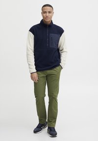 Pull en polaire marine avec des manches crème, fermeture éclair quart et poche poitrine. Associé à un pantalon vert olive et des baskets bleu foncé.
