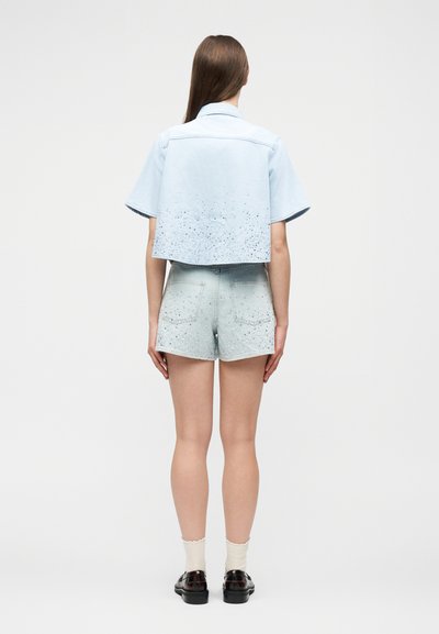 Γυναίκα με μακριά καστανά μαλλιά που στέκεται γυρισμένη προς τα πίσω, φοράει γαλάζιο κοντομάνικο πουκάμισο από τζιν και τζιν σορτς ψηλόμεσο με μαύρα πιτσιλίσματα.