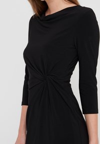 Kobieta w czarnej sukience z długim rękawem, z węzłem na talii, odsłaniająca górną część ciała i ramiona.