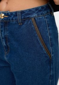 Denim traperice tamnoplave boje s zlatnim dugmadima, sa kontrastnim smešnim šavovima i detaljem bočne džepa.