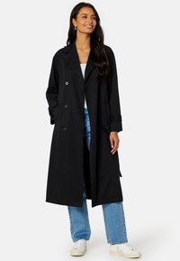 Černý dlouhý trench coat s dvojitým zapínáním, velkými knoflíky, širokým límcem a ležérním střihem, nošený přes modré džíny a tenisky.