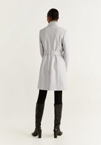 Manteau gris ceinturé au design ajusté, confectionné dans un mélange de laine douce. Il présente un col montant et une longueur mi-cuisse, assorti à de hautes bottes noires.