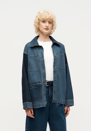 Personne aux cheveux blonds bouclés portant une veste en jean bicolore sur une chemise blanche et un pantalon en jean bleu foncé avec une ceinture noire.