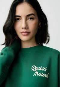 Grøn sweatshirt med rund halsudskæring og en blød tekstur. Den har teksten "Rockin' Around" i lys creme farve broderet på brystet.