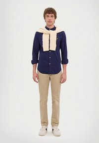 Μπλε ναυτικό πουκάμισο με κουμπιά, κρεμ πουλόβερ από πλεκτό ρίχνεται πάνω, μπεζ παντελόνι chino, λευκά αθλητικά παπούτσια με μαύρες λεπτομέρειες, στέκεται μπροστά σε ουδέτερο φόντο.