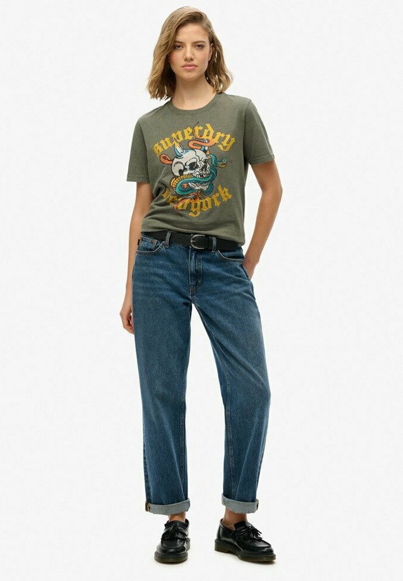 Camiseta gráfica verde oliva con un diseño de calavera y serpiente, combinada con jeans de tiro alto en azul y zapatos negros con cordones.