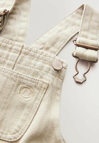 Salopette en denim beige avec un tissu texturé, dotée de bretelles ajustables, de matériel en bronze, d'un patch logo circulaire et d'une poche avant.