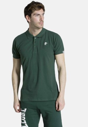 Uomo che indossa una polo verde scuro e pantaloni abbinati con scritta bianca, guarda di lato su sfondo semplice.