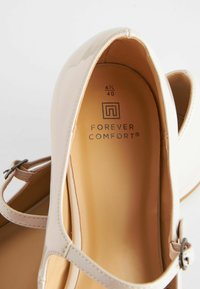 Chaussure en cuir verni beige avec un bout arrondi, une bride de cheville dotée d'une boucle en argent et un intérieur lisse et rembourré de couleur beige.