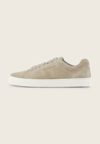 Zapatilla de ante beige con cordones gris claro, suela de goma blanca y un sutil detalle del logo. Textura suave y diseño aerodinámico para mayor comodidad.