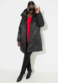 Giacca a vento nera con cappuccio, dotata di chiusura a zip e bottoni. Indossata sopra un top rosso, abbinata a pantaloni skinny neri e tacchi a punta neri.
