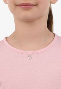 Collar de cadena de plata con un colgante redondo de diseño de corona y un pequeño acento rosa, llevado sobre una camisa de color rosa claro.