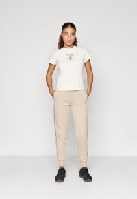 Κρέμα κοντομάνικο μπλουζάκι με το λογότυπο "GUESS", συνδυασμένο με μπεζ φόρμες που διαθέτουν πλαϊνές ρίγες, και μαύρα αθλητικά παπούτσια. Λείες υφασμάτινες υφές.