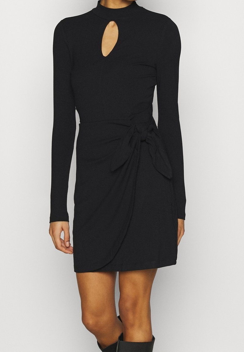 Femme portant une mini-robe noire à manches longues avec un décolleté en forme de trou de serrure et un détail noué à la taille, associée à des bottes noires.