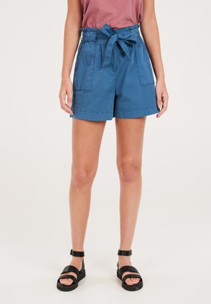 Blaue Baumwollshorts mit schmalem Bund, Bindegürtel, zwei seitlichen Taschen und umgeschlagenem Saum. Getragen mit schwarzen, riemchenförmigen Sandalen.