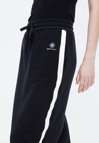 Pantaloni sportivi neri con strisce laterali bianche, dotati di una vita con cordoncino e piccolo logo ricamato sulla coscia sinistra. Tessuto morbido.