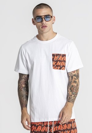 Hombre con tatuajes que lleva gafas de sol azules, camiseta blanca con bolsillo de estampado naranja y pantalones cortos a juego con el mismo estampado naranja.