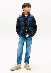 Marinblå vadderad väst över en rutig skjorta, kombinerad med blekta blå jeans och beiga sneakers. Vänsten har sidfickor och dragkedja.