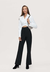 Blazer blanco recortado con mangas largas, combinado con pantalones negros de cintura alta. La modelo sostiene un bolso a cuadros y lleva botas negras hasta el tobillo.