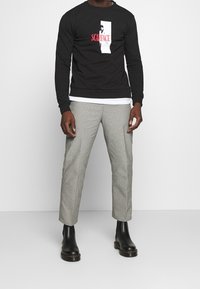 Sudadera negra con impresión gráfica de "Scarface", combinada con pantalones gris claro de corte ajustado y botas negras de tobillo.
