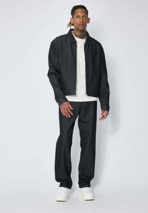 Homme avec des tatouages et des dreadlocks portant une veste et un pantalon sombres avec une chemise blanche et des baskets blanches, debout devant un fond uni.