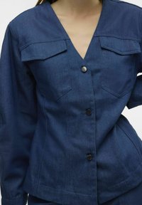Giubbotto in denim blu scuro, con scollatura a V, due tasche sul petto, bottoni neri e una vestibilità strutturata con una lunghezza leggermente accorciata.