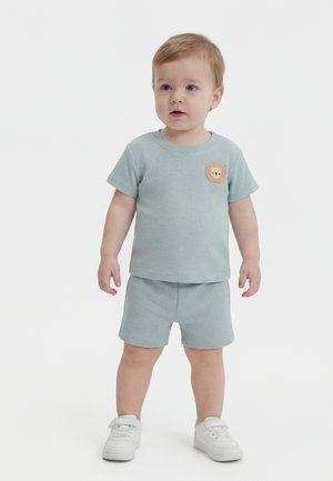 Παιδάκι όρθιο, φορώντας ανοιχτό μπλε κοντομάνικη μπλούζα και σορτς με κεντημένο λιοντάρι, και λευκά αθλητικά παπούτσια σε απλό λευκό φόντο.