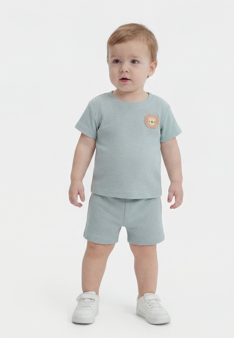 Παιδάκι όρθιο, φορώντας ανοιχτό μπλε κοντομάνικη μπλούζα και σορτς με κεντημένο λιοντάρι, και λευκά αθλητικά παπούτσια σε απλό λευκό φόντο.