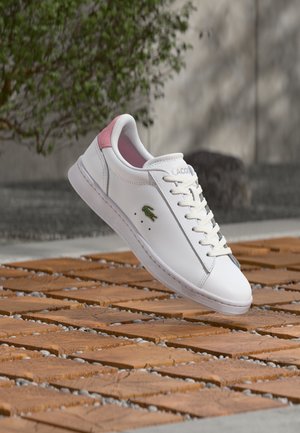 Biały skórzany sneaker z różowym detalem na pięcie i zielonym logo krokodyla, unoszący się nad drewnianymi płytkami z rozsypanymi kamykami na zewnątrz.