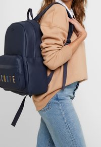 Nainen, joka k äyttää beigeä neuletta ja sinisiä farkkuja, ja hänen olkapäänsä yli on navy sininen Lacoste-reppu valkoista taustaa vasten.