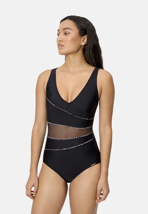 Mujer con traje de baño de una pieza negro con paneles transparentes en la sección media y líneas diagonales blancas con patrones, sobre un fondo claro y liso.