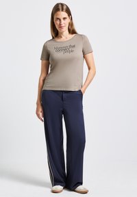 Camiseta color caqui con texto negro, combinada con pantalones de pierna ancha en color marino con una franja dorada a los lados. El modelo lleva zapatos de color claro.