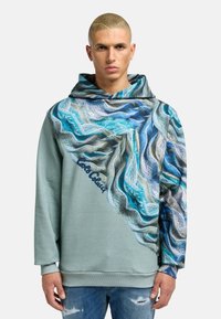 Fleece dukserica s kapuljačom u svijetlozelenoj boji s teksturiranim plavim valovitim uzorkom. Ima veliku kapuljaču, rebraste manžete i izvezeni logo u tamnoplavoj boji.
