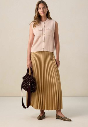 Femme portant un gilet beige sans manches à boutons, une jupe maxi plissée dorée, des ballerines en serpent, et tenant un sac à main rouge foncé avec des œillets.
