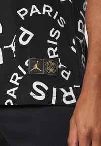 Czarna tkanina z białym napisem "PARIS" oraz logosami Jumpman, z małą czarną i złotą naszywką z symbolami Paris Saint-Germain i Jumpman.