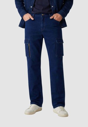 Uomo che indossa pantaloni cargo blu scuro con tasche con zip e patta, sneaker bianche e giacca blu scuro con le mani nelle tasche.