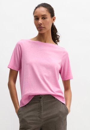 Donna con capelli ricci che indossa una camicia rosa chiaro a maniche corte infilata parzialmente in pantaloni grigio scuro, mani nelle tasche, sfondo neutro.