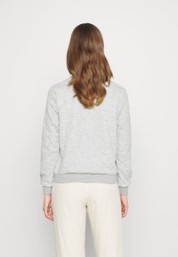 Sudadera gris claro con tejido texturizado, puños y bajo acanalados. La parte trasera muestra un diseño minimalista, combinado con pantalones de color crema.