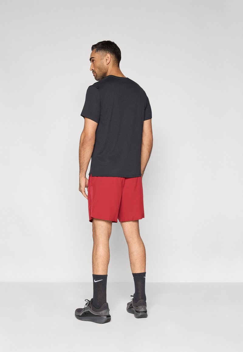 T-shirt nera a maniche corte abbinata a pantaloncini rossi. Il modello indossa calzini scuri e scarpe da ginnastica nere. Design semplice e aderente, texture liscia.