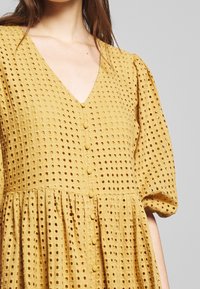 Senapsgul klänning med en texturerad, perforerad tyg. Har en V-ringning, puffärmar och knappar längs framsidan.