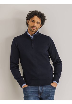 Marinefarbener Reißverschluss-Pullover mit gerippten Bündchen und Saum, aus Strickstoff gefertigt. Über einem hellblauen Hemd mit Kragen getragen, kombiniert mit Denim-Jeans.
