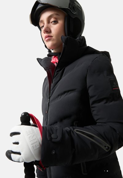 Schwarze gepolsterte Skijacke mit hohem Kragen, Reißverschluss und Knopfverschluss sowie grauen Akzent-Reißverschlüssen; getragen mit weißen Handschuhen, die einen Skistock halten.