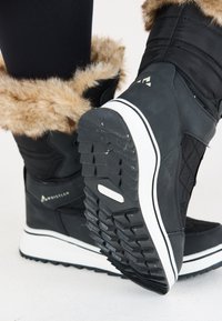 Svart vinterkänga med ett fuskpälskant, quiltad textur, vit mellansula och mönstrad gummisula för grepp. Logotyp synlig på sidan.