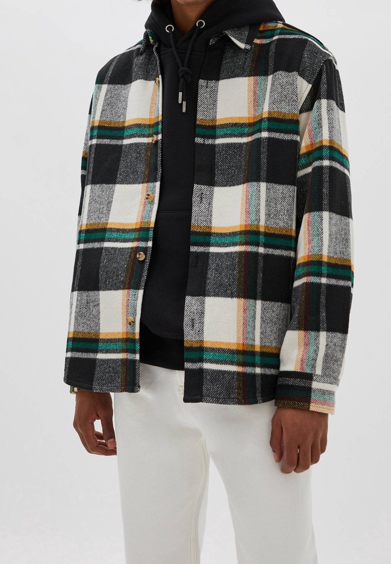 Homme portant un sweat à capuche noir sous une chemise à carreaux noire, blanche et verte avec un pantalon blanc sur un fond uni.