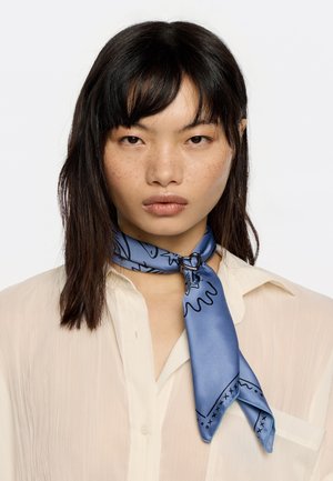 Silk scarf in light blue, featuring black abstract patterns. Tied around the neck with a heart-shaped silver clasp, paired with a cream shirt.