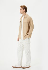 Veste en tissu texturé beige avec deux poches poitrine, associée à un pantalon cargo blanc et des baskets blanches avec des accents marron.
