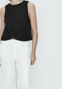 Haut noir sans manches avec un design frontal torsadé ; associé à un jean blanc taille haute. Le haut a une texture côtelée.