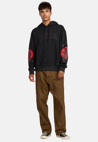 Schwarzer Hoodie mit RVCA-Logo, roten Tigergrafiken auf den Ärmeln, einer Fronttasche und Kapuze mit Kordelzug. Kombiniert mit braunen Hosen und schwarzen Sneakern.