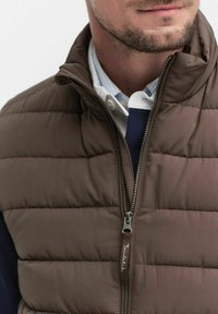 Brązowa pikowana kamizelka z wysokim kołnierzem i zapięciem na zamek błyskawiczny. Posiada przeszywane panele i widoczny metkę marki na uchwycie zamka.